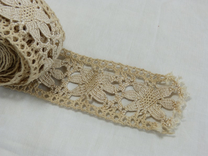 May include: A roll of beige lace with a floral design. The lace is made of a woven material and has a delicate, openwork pattern. The edge of the lace is fringed. The lace is likely used for sewing, crafting, or adding a decorative touch to garments.