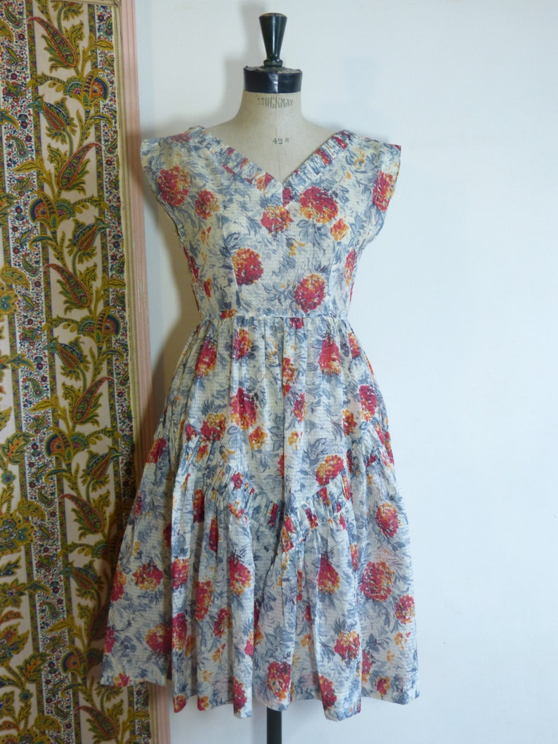 May include: A vintage sleeveless dress with a V-neckline and a floral print in shades of red, yellow, and blue on a white background. The dress has a tiered skirt and a fitted bodice.