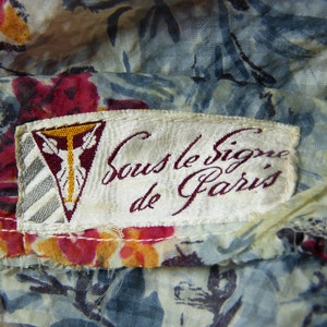 May include: A white fabric label with the text "Sous le Signe de Paris" in burgundy cursive lettering. The label has a small, gold-coloured triangle with a stylized figure in the centre.