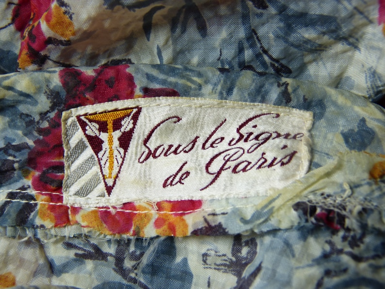 May include: A white fabric label with the text "Sous le Signe de Paris" in burgundy cursive lettering. The label has a small, gold-coloured triangle with a stylized figure in the centre.