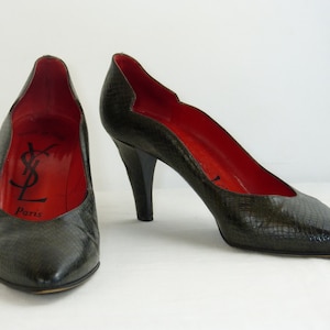 Peut inclure: Une paire de escarpins à talons en peau de serpent verte avec une doublure en cuir rouge. Les chaussures ont une pointe pointue et un talon aiguille haut. L'intérieur de la chaussure porte le logo Yves Saint Laurent et le mot "Paris" imprimé en noir.