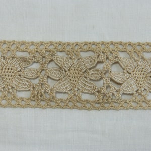 May include: Close-up of a beige crocheted lace trim with a floral design. The trim features a repeating pattern of stylized flowers and openwork details, suitable for various craft projects.