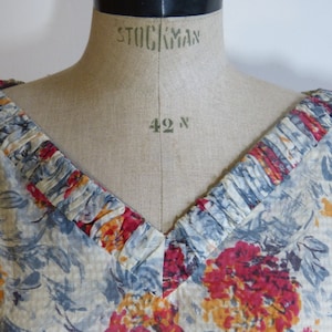 May include: A white dress with a blue and red floral print. The neckline has a gathered ruffle detail.