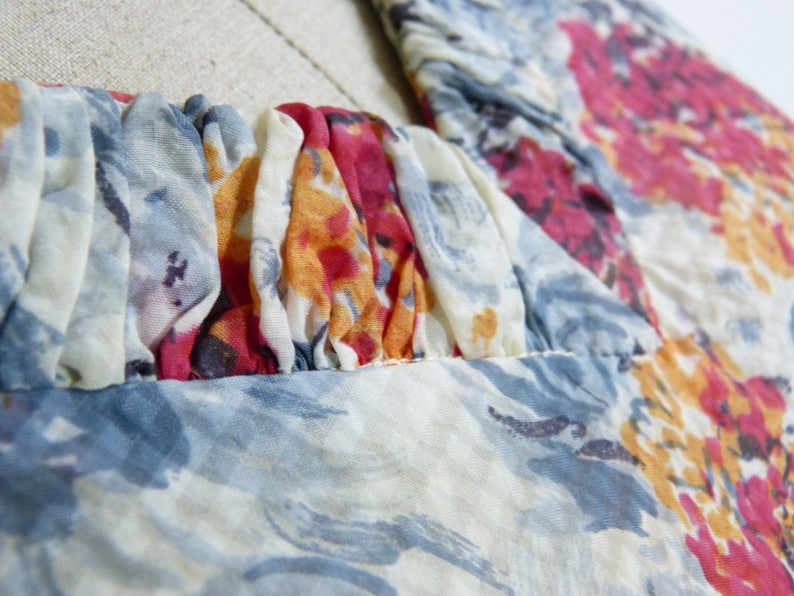 May include: Close-up of a white, blue and pink floral print fabric with a gathered neckline. The fabric is sheer and has a delicate texture.