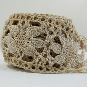 May include: A beige crocheted lace trim with a floral design. The trim features an openwork pattern with leaf-shaped details and fringe along one edge. This decorative trim is suitable for various craft projects.