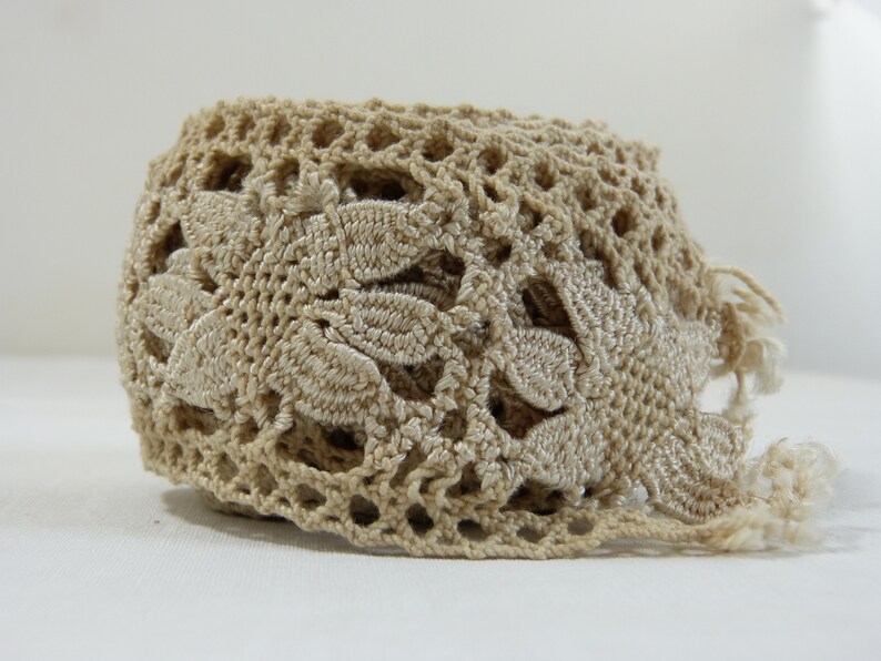 May include: A beige crocheted lace trim with a floral design. The trim features an openwork pattern with leaf-shaped details and fringe along one edge. This decorative trim is suitable for various craft projects.