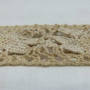 May include: Close-up of a beige crocheted lace trim. The trim features a repeating floral pattern with openwork details. The lace is made of a soft, textured yarn and is set against a white background.