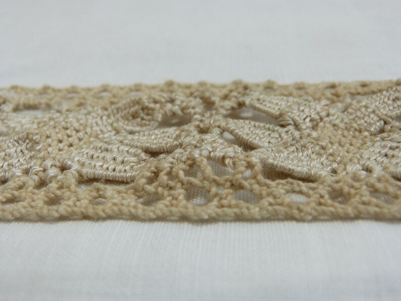 May include: Close-up of a beige crocheted lace trim. The trim features a repeating floral pattern with openwork details. The lace is made of a soft, textured yarn and is set against a white background.