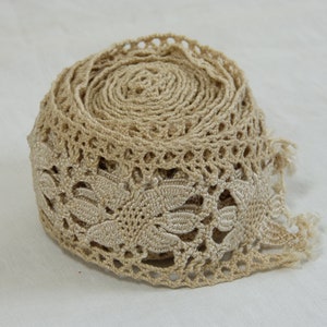 May include: A roll of beige lace trim with a floral leaf design. The trim is made of a woven material and has a delicate, openwork pattern. The edge of the trim has a scalloped design and a fringe.