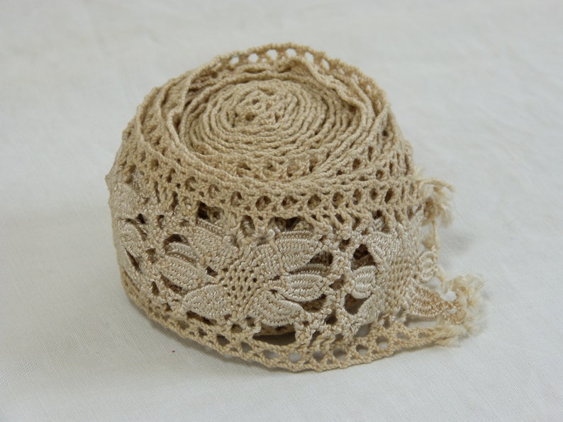 May include: A roll of beige lace trim with a floral leaf design. The trim is made of a woven material and has a delicate, openwork pattern. The edge of the trim has a scalloped design and a fringe.