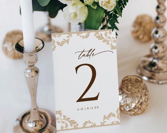 Könnte beinhalten: Eine Tischnummerkarte mit floralem Design und der Zahl 2 in Braun. Die Karte liegt auf einer weißen Tischdecke, neben einem silbernen Kerzenständer und einem Blumengesteck mit weißen Blumen. Die Karte lautet "Table 2".