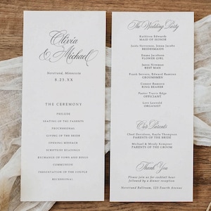 May include: Two white wedding programs with elegant calligraphy, featuring the names "Olivia & Michael" and details of the ceremony and wedding party. The programs are set on a wooden surface with a white fabric and dried flowers.