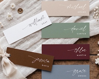 May include: A set of rectangular paper place cards in white, beige, sage green, burgundy, brown, and slate blue. Each card features a name printed in a white or black script font with the surname in a clean, sans-serif capital letter font.