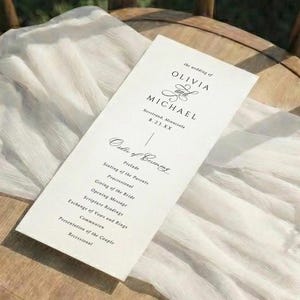 May include: A wooden chair with a round seat holds a white fabric and a wedding program. The program, with the names "Olivia & Michael", lists the order of events. The scene is set outdoors with greenery and sunlight.