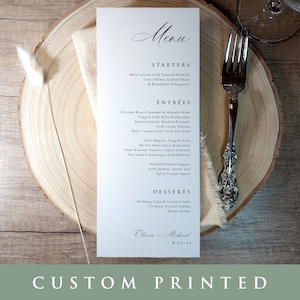 May include: A white wedding menu card with a black script font. The menu includes starters, entrees, and desserts. The card is on a wooden surface with a white napkin and a silver fork.