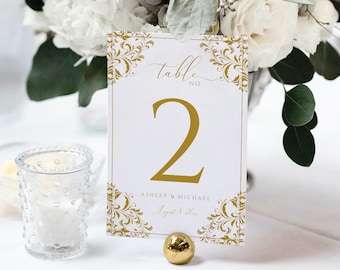 Könnte beinhalten: Goldene Tischnummerkarte mit weißem Hintergrund und verziertem goldenen Rand. Die Karte trägt die Aufschrift "Table No. 2 Ashley & Michael August 5, 2023".