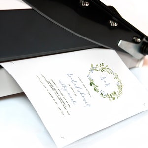 May include: A white card with a blue and green floral wreath with the initials "A+K" in the center. The text "Please join us for a bridal shower" is written in cursive above the wreath. The text "In honor of Ashley Winch" is written below the wreath. The text "Saturday, August 5th at 3pm" is written below the text "In honor of Ashley Winch". The text "Janice Sower's Residence" is written below the date. The text "123 Fourth Street, Intertrand, Minnesota" is written below the residence. The text "Please RSVP to Janice at 307-535-1212" is written below the address.