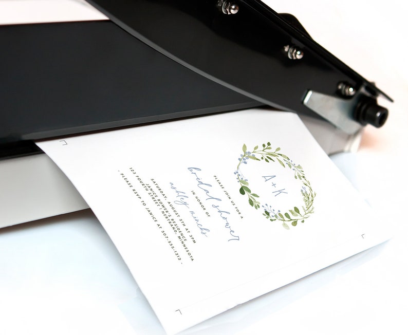 May include: A white card with a blue and green floral wreath with the initials "A+K" in the center. The text "Please join us for a bridal shower" is written in cursive above the wreath. The text "In honor of Ashley Winch" is written below the wreath. The text "Saturday, August 5th at 3pm" is written below the text "In honor of Ashley Winch". The text "Janice Sower's Residence" is written below the date. The text "123 Fourth Street, Intertrand, Minnesota" is written below the residence. The text "Please RSVP to Janice at 307-535-1212" is written below the address.