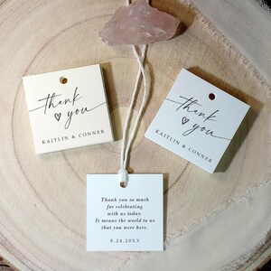 Op de afbeelding: Twee witte bedankkaartjes met zwarte kalligrafie die "Bedankt" met een hartje zegt. De kaartjes zijn aan elkaar gebonden met een wit touwtje en hebben de namen "Kaitlin & Conner" erop gedrukt. Het derde kaartje is wit met zwarte tekst die zegt "Hartelijk bedankt voor het vieren met ons vandaag. Het betekent de wereld voor ons dat je er was. 8.24.20XX".