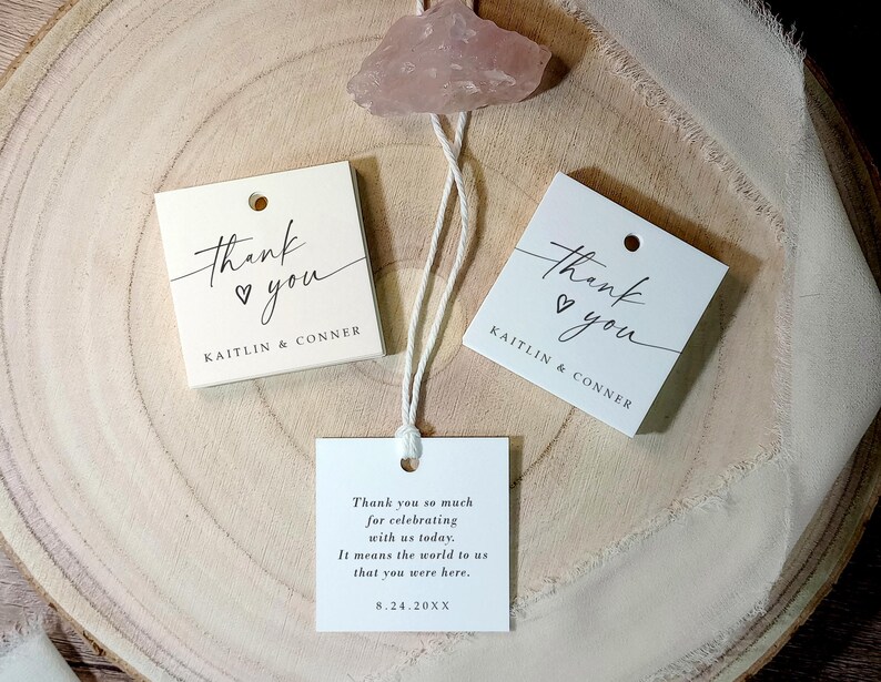 Op de afbeelding: Twee witte bedankkaartjes met zwarte kalligrafie die "Bedankt" met een hartje zegt. De kaartjes zijn aan elkaar gebonden met een wit touwtje en hebben de namen "Kaitlin & Conner" erop gedrukt. Het derde kaartje is wit met zwarte tekst die zegt "Hartelijk bedankt voor het vieren met ons vandaag. Het betekent de wereld voor ons dat je er was. 8.24.20XX".