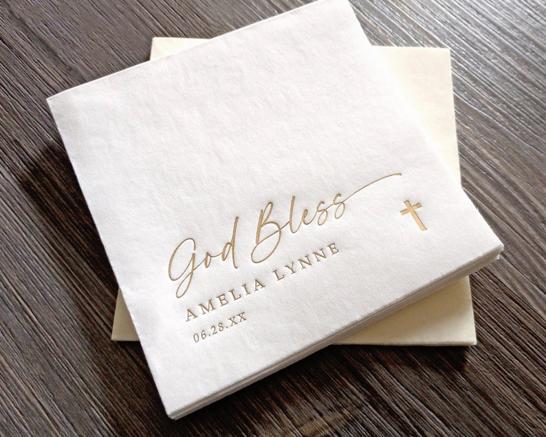 May include: White paper napkins with gold foil lettering that reads "God Bless Amelia Lynne 06.28.XX" and a gold cross.
