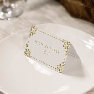 May include: A white place card with a gold ornate border. The text on the place card reads 'Michael Baker, Table 2.'