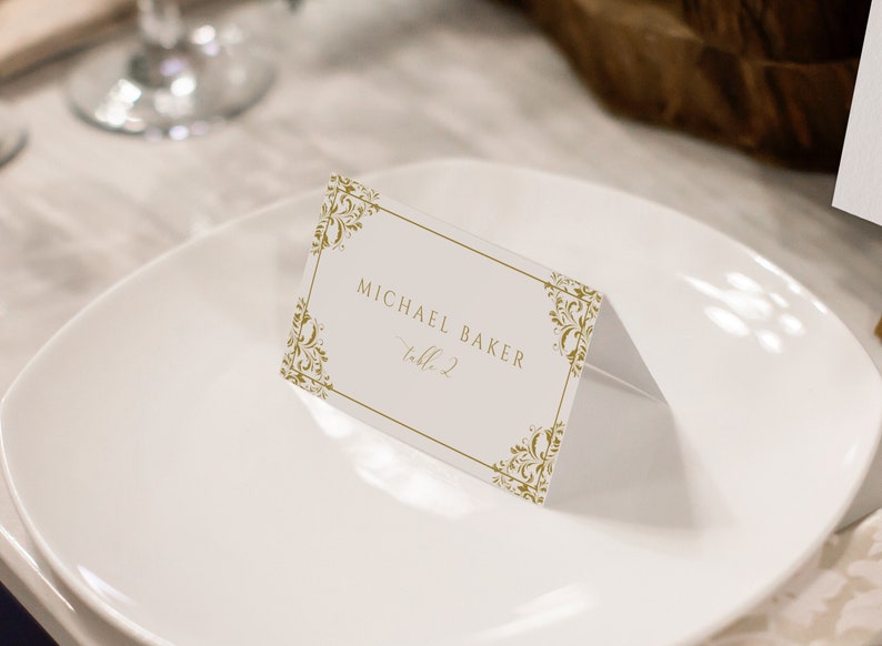May include: A white place card with a gold ornate border. The text on the place card reads 'Michael Baker, Table 2.'