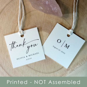 Op de afbeelding: Twee witte vierkante bruiloft-favorieten-labels met touw. Een label zegt "dank je" met de namen Olivia & Michael en de datum 8.23.XX. Het andere label heeft de initialen O|M en de datum 23 augustus 20XX.