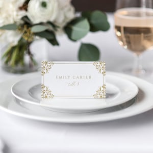 May include: A white place card with a gold ornate border. The text on the card reads 'Emily Carter, table 8.'