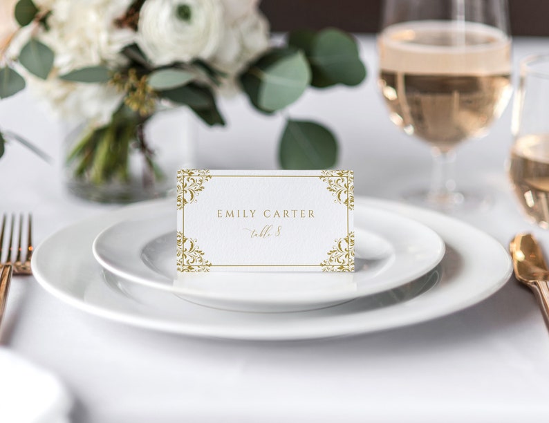 May include: A white place card with a gold ornate border. The text on the card reads 'Emily Carter, table 8.'