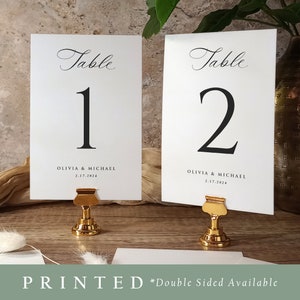 May include: Two white table number cards with black calligraphy. The cards are on gold metal stands with a clip to hold the cards. The cards are numbered 1 and 2. The text on the cards reads "Table" and "Olivia & Michael 2.17.2024".