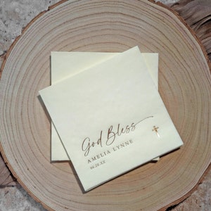 May include: A white card with the text "God Bless Amelia Lynne 06.28.XX" in brown script. A small gold cross is to the right of the text.