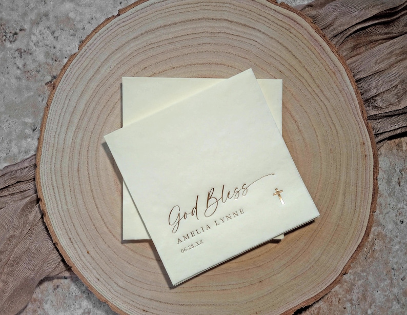 May include: A white card with the text "God Bless Amelia Lynne 06.28.XX" in brown script. A small gold cross is to the right of the text.