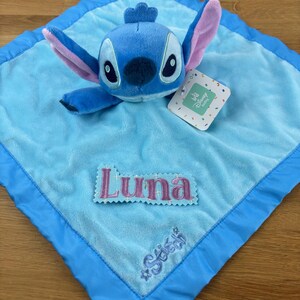 May include: A blue plush Stitch character toy rests on a light blue baby blanket. The blanket has a darker blue border and features the name "Luna" embroidered in pink, along with the name "Stitch". A Disney Baby tag is attached.