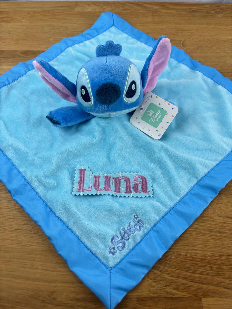 May include: A blue plush Stitch character toy rests on a light blue baby blanket. The blanket has a darker blue border and features the name "Luna" embroidered in pink, along with the name "Stitch". A Disney Baby tag is attached.