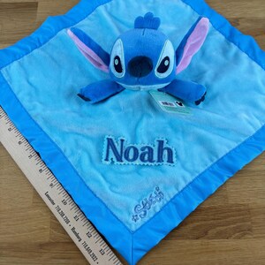 May include: A blue plush baby blanket featuring a Stitch character. The blanket has a light blue center with a darker blue border. The name "Noah" is embroidered on the blanket, along with the hashtag "#Stitch".