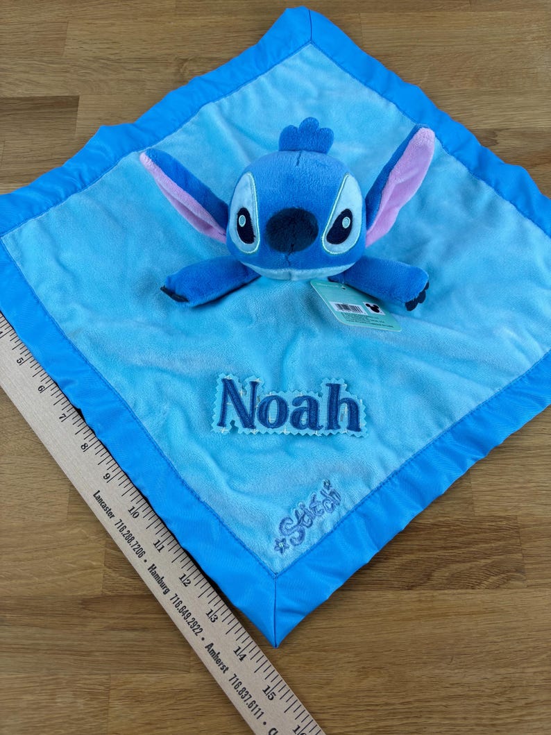 May include: A blue plush baby blanket featuring a Stitch character. The blanket has a light blue center with a darker blue border. The name "Noah" is embroidered on the blanket, along with the hashtag "#Stitch".