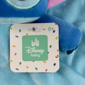 May include: A blue plush toy with a tag that reads "Disney baby". The tag has a green square with a castle logo and the words "Disney baby" in white. The tag also has colorful confetti-like shapes around the edges.