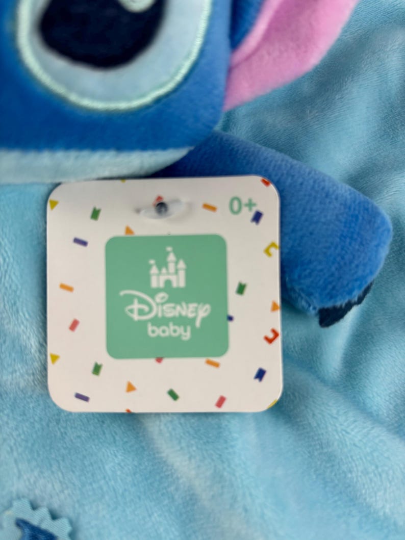 May include: A blue plush toy with a tag that reads "Disney baby". The tag has a green square with a castle logo and the words "Disney baby" in white. The tag also has colorful confetti-like shapes around the edges.