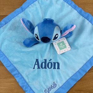 May include: A blue plush baby blanket featuring a Stitch character with a matching blue border. The blanket has the name "Ad&oacute;n" embroidered in blue, along with the Stitch logo. A Disney baby tag is attached.