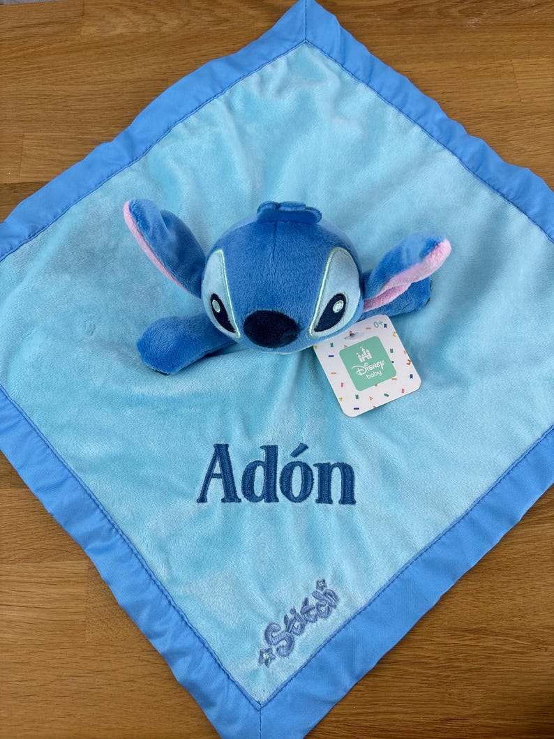 May include: A blue plush baby blanket featuring a Stitch character with a matching blue border. The blanket has the name "Ad&oacute;n" embroidered in blue, along with the Stitch logo. A Disney baby tag is attached.