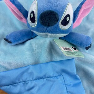 May include: A blue plush Stitch toy with large ears and a dark blue nose, resting on a light blue blanket. The blanket has a satin border and a tag attached. The toy is a character from the Disney movie Lilo & Stitch.