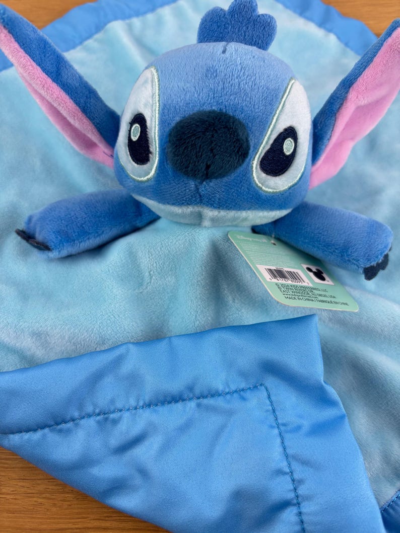 May include: A blue plush Stitch toy with large ears and a dark blue nose, resting on a light blue blanket. The blanket has a satin border and a tag attached. The toy is a character from the Disney movie Lilo & Stitch.