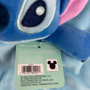 May include: A close-up of a blue plush toy of the character Stitch from Disney's Lilo & Stitch, with pink ears and a tag. The tag includes the Disney Baby logo, a barcode, and the Kids Preferred LLC address.