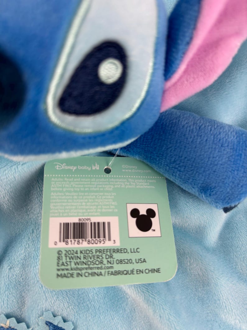 May include: A close-up of a blue plush toy of the character Stitch from Disney's Lilo & Stitch, with pink ears and a tag. The tag includes the Disney Baby logo, a barcode, and the Kids Preferred LLC address.
