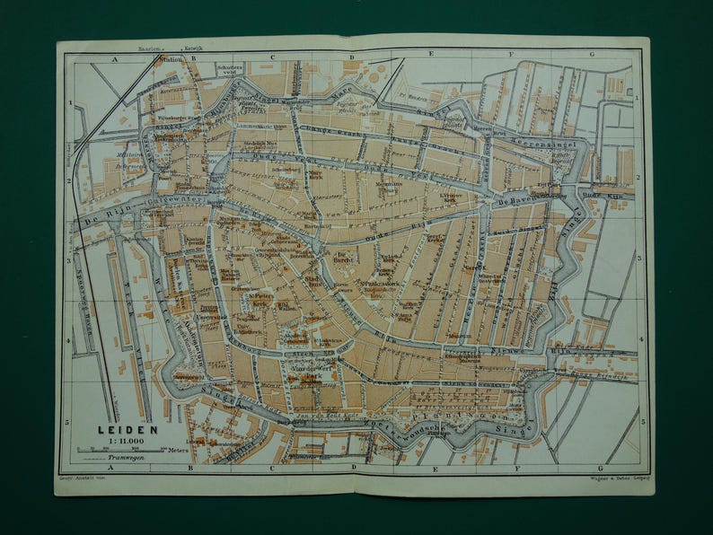 May include: A vintage map of Leiden, Netherlands, featuring a detailed street layout with canals and buildings. The map is printed in sepia tones with black and gray details. The title "LEIDEN" is visible at the bottom, along with a scale of 1:11,000.