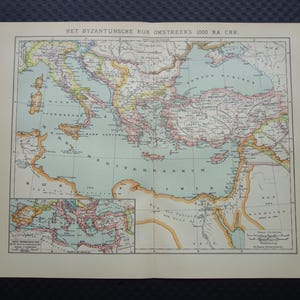 Op de afbeelding: Een vintage kaart van het Byzantijnse Rijk rond 1000 na Christus, met gedetailleerde geografische kenmerken en tekst in het Nederlands. De kaart heeft een hoofdonderdeel en een inzetkaart, met een kleurenpalet van beige, blauw, groen en roze.