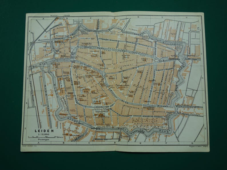 May include: An open vintage map of Leiden, Netherlands, with detailed streets, canals, and buildings in brown and gray tones. The map is printed on aged paper, with the city name and scale visible.