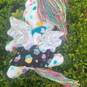 May include: A white plush toy with a rainbow yarn tail and wings. The toy has a polka dot pattern and a cupcake-themed fabric skirt.