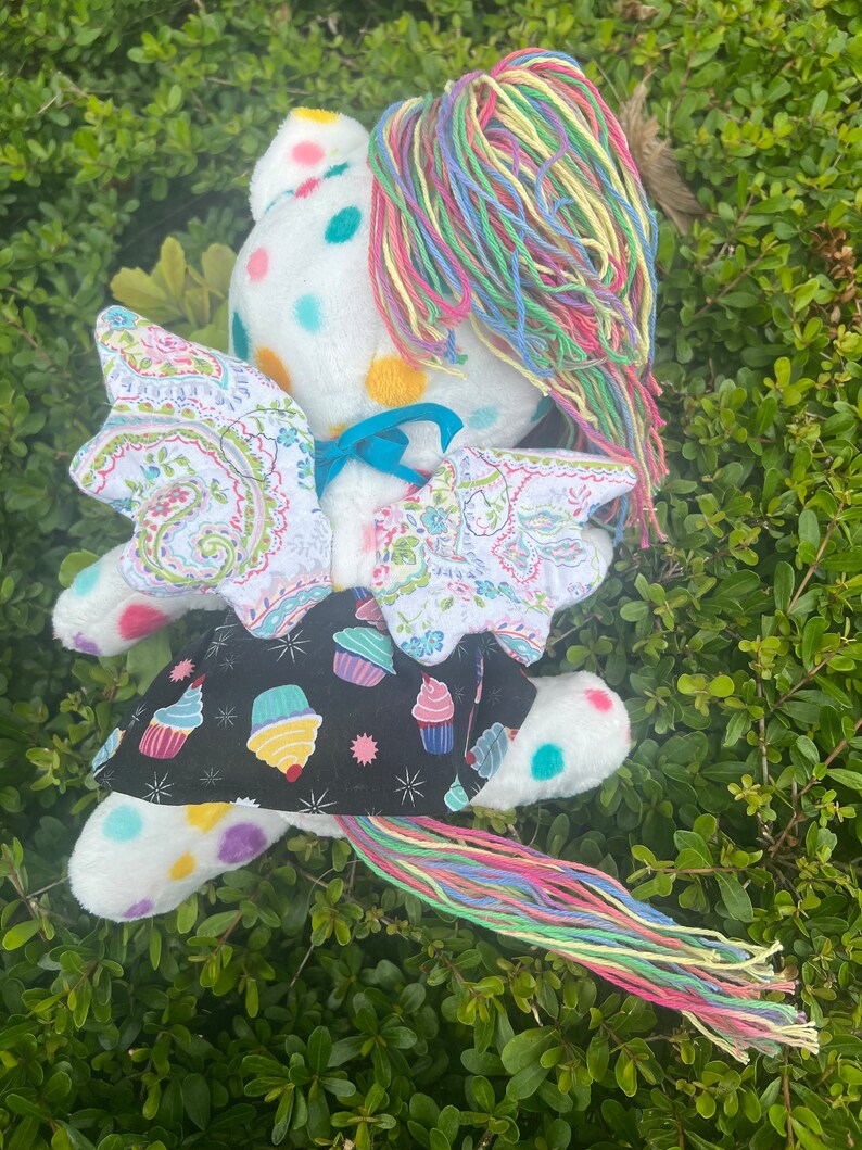 May include: A white plush toy with a rainbow yarn tail and wings. The toy has a polka dot pattern and a cupcake-themed fabric skirt.